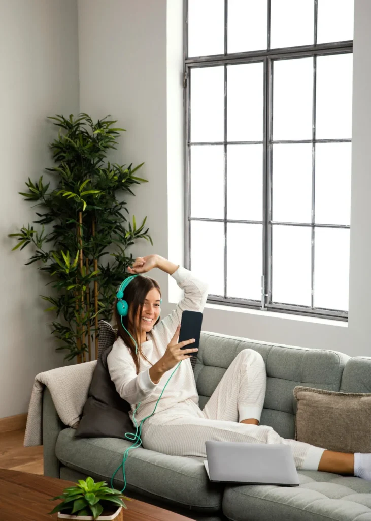 Young woman in pajamas smiling while taking a selfie during a video call, wearing teal headphones and lounging on a sofa with a laptop beside her.