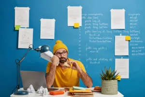 Frustrated programmer in yellow outfit holding sandwich and crumpled paper, sitting at a cluttered desk with laptop and coding notes on a blue wall.