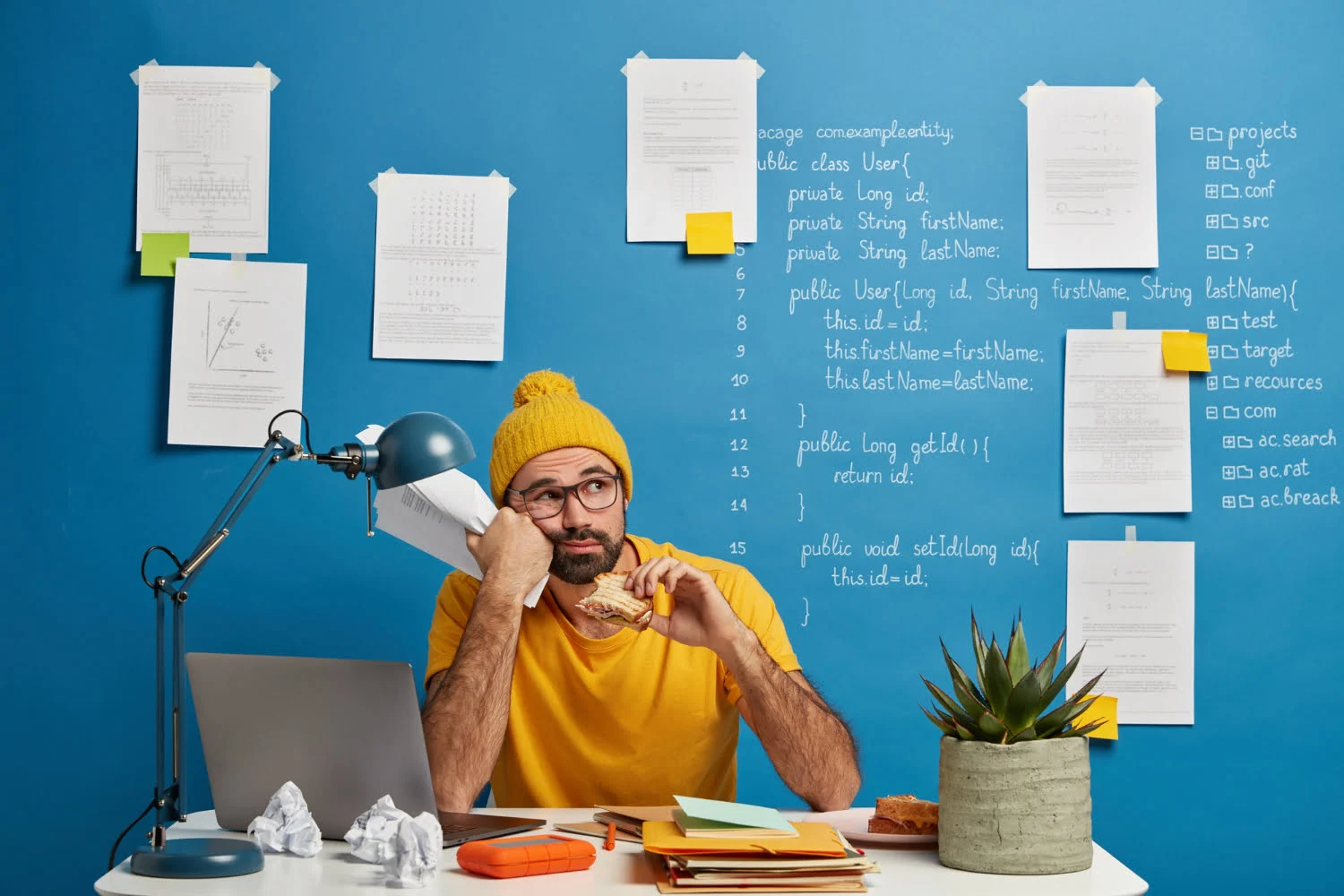 Frustrated programmer in yellow outfit holding sandwich and crumpled paper, sitting at a cluttered desk with laptop and coding notes on a blue wall.